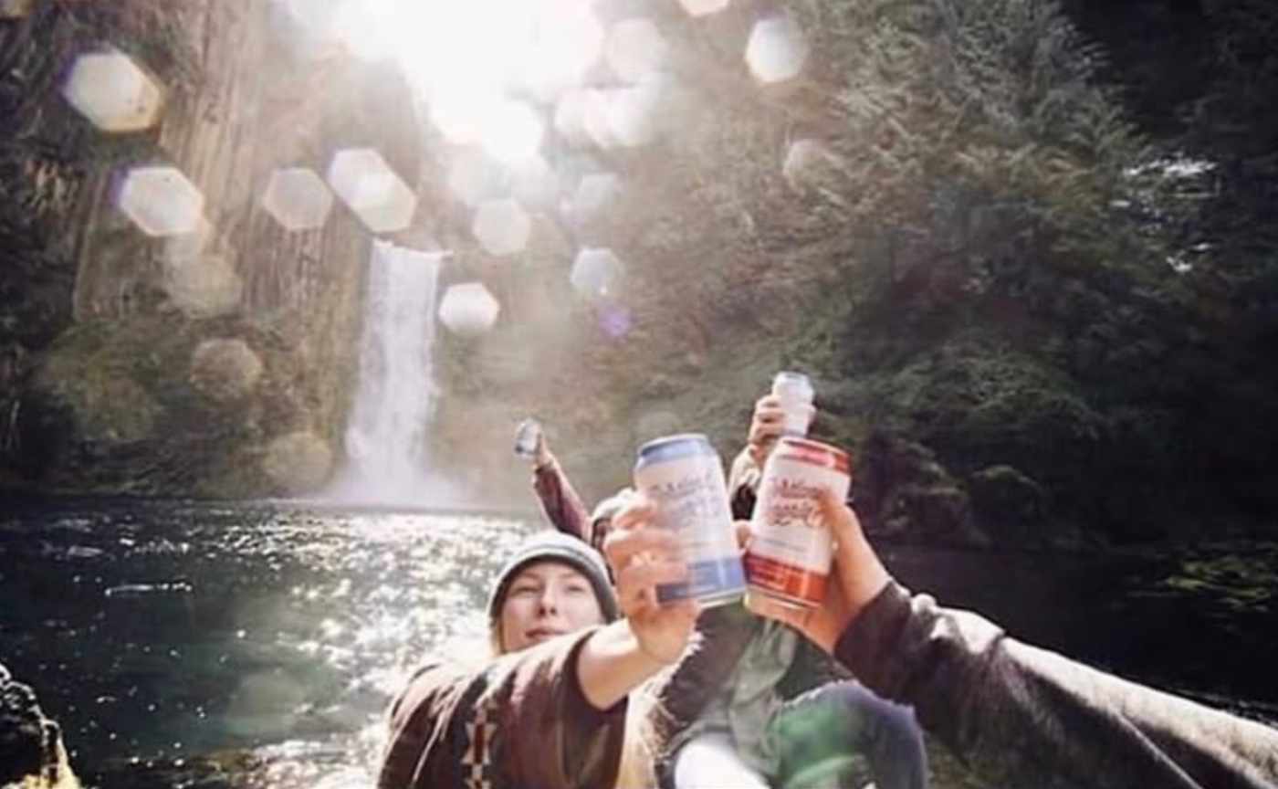 Two friends toasting cans of Portland Sangria beneath a Pacific Northwest waterfall